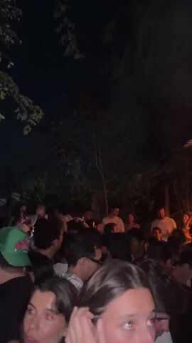 The video shows a crowd of people at an outdoor party or club at night. The camera moves through the crowd, showing faces and the structure overhead which is lit with orange and blue lights. Some of the people are wearing light-colored shirts. Trees and foliage are visible in the background. The music is loud.