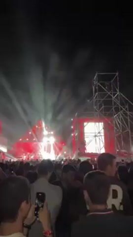 The video shows a dark, outdoor scene of a large crowd gathered in front of a stage. The stage is illuminated with bright white and red lights, and a large screen on the right side displays a performer. A metal structure surrounds the screen. Many people in the crowd are holding up their phones, presumably to record the event. The crowd appears dense, with people standing close together. The overall atmosphere seems energetic and celebratory, likely a concert or festival.