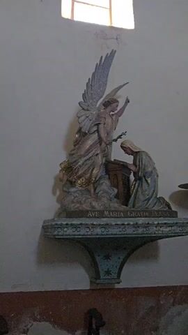 The video pans across the interior of a church, showcasing various religious artifacts and architectural elements. Initially, a statue of an angel is visible, positioned above a pedestal inscribed with "AVE MARIA GRATIA PLENA". The camera then moves to a framed relief depicting a biblical scene with multiple figures, followed by a large painting situated above an altar. The altar itself is adorned with candles and smaller statues. The scene also includes ornate columns, a decorative railing, and a window allowing natural light to enter the space. The overall impression is one of a richly decorated and historical place of worship.