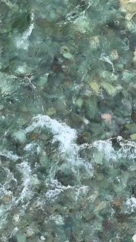 The video shows a top-down view of a shallow stream with clear, greenish water flowing over rocks. The sunlight creates patterns on the water and illuminates the rocks beneath. The water is slightly turbulent, with small ripples and occasional white foam.