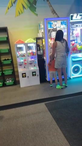 The video shows a woman and a small child standing in front of two arcade claw machines. The woman is wearing a white tank top and denim shorts, and the child is wearing an orange t-shirt and gray shorts. Both are wearing bright green socks. The claw machines are filled with colorful stuffed animals. The machine on the right is labeled "PRIZE CUBE" and has blue and purple lights. The machines on the left are smaller and appear to be for dispensing smaller prizes. The background shows a tropical-themed mural with a palm tree. The floor is carpeted. The child is looking at the prizes in the machine on the right. The woman has her arm around the child.