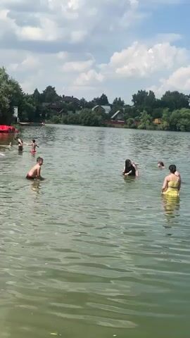 The video depicts a sunny day at a lake or reservoir, likely a recreational area. The water is calm and appears to be a popular spot for swimming and relaxation. Several people are visible in the water, including children and adults, engaged in various activities. One boy is standing in shallow water, appearing to interact with someone off-camera. A woman in a yellow swimsuit is also in the water, as are a couple of other individuals. The shoreline features a sandy beach with lounge chairs and umbrellas, suggesting a developed public beach. Trees and houses are visible in the background, indicating a semi-rural or suburban setting. The sky is blue with scattered white clouds, characteristic of a pleasant summer day. The overall atmosphere appears to be one of leisure and enjoyment. There is no visible text in the video.