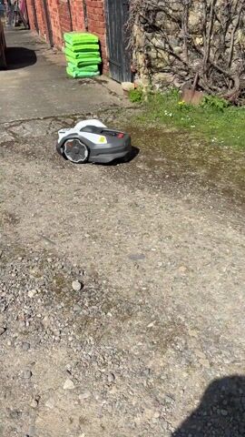 The video shows a robot lawnmower sitting on a gravel surface. The mower is white and gray with red accents and large, patterned wheels. Behind the mower is a concrete driveway or path, a dark-colored car, several green bags stacked against a red brick building, and a black wooden door set into a stone wall with some vegetation growing on it. The ground is a mix of gravel and concrete, with some damp or wet patches visible. The sun appears to be out, casting a shadow in the foreground. The camera remains still throughout the video.