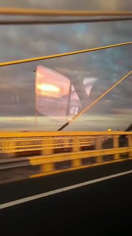 The video is a first-person perspective of traveling across a bridge during sunset. The camera appears to be mounted on a vehicle. The sky is filled with dramatic clouds, illuminated by the setting sun, casting a warm glow on the scene. The bridge itself is a cable-stayed bridge with prominent yellow support beams and cables. The road surface is visible, and there are lane markings. Reflections of the sky and the bridge's structure are visible in what appears to be the vehicle's windows, creating a layered and somewhat abstract visual effect. The movement of the vehicle is evident through the shifting perspective and the motion blur in the background. No people or specific interactions are clearly visible.