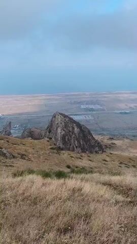 The video begins with a view of a rocky, grassy landscape with a large rock formation in the foreground. The camera pans to the left, revealing a valley with a body of water in the distance and some buildings. The video continues to pan left, showing more of the rocky terrain and a glimpse of the distant water. The camera then moves upwards, focusing on a large, jagged rock formation that dominates the frame. The video ends with a shot of the rocky landscape with the distant water and sky visible.