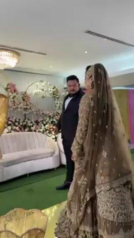 The video shows a wedding scene. A bride in a brown embroidered dress and veil stands beside a man in a black suit. They are both looking towards the side, possibly at guests or an event taking place. The backdrop includes floral decorations and a white couch. A large gold decorative vase is in the foreground, partially obscuring the view. The lighting is bright and the flooring appears to be green carpet. The location seems to be an indoor event hall. There is music playing in the background.