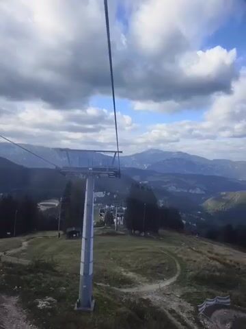 The video shows a view from a ski lift ascending a mountain. A ski lift tower is visible in the foreground, with the cable extending upwards. The mountain landscape unfolds in the background, with trees, a small lake, and distant mountain ranges under a cloudy sky. The ski slope below is covered in grass, indicating it is not winter.
