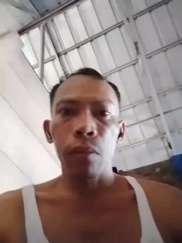The video shows a close-up, low-angle shot of a man's face. He is wearing a white tank top. His skin has some blemishes and uneven tone. He has short, dark hair, and is wearing small, dark earbuds in both ears. The background appears to be a room with a corrugated metal ceiling and some white walls. There is also a blue tarp visible in the upper right corner. The man's mouth is slightly open and he appears to be speaking, but his words are unclear due to audio quality. He blinks and shifts his gaze slightly during the short clip.