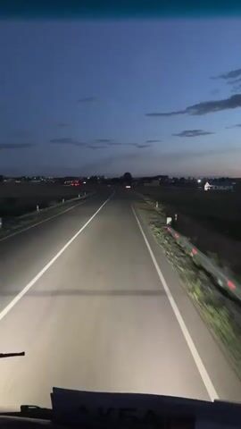 This video captures a first-person perspective of driving on a road in Baiqadam, Kazakhstan, during twilight. The scene is dominated by the asphalt road stretching forward, illuminated by the vehicle's headlights, with a clear white line marking the center. To the sides of the road, the landscape consists of flat terrain with fields and scattered buildings, some of which appear to be houses or farm structures. In the distance, faint lights suggest a settlement or ongoing activity. The sky is a deep blue, transitioning from lighter hues near the horizon, with a few wispy clouds adding texture. The overall mood is serene and somewhat solitary, typical of a journey during the fading light of day. No people are directly visible in the frame, and the focus is on the act of driving and the evolving environment. The time of day is clearly evening, as indicated by the low light and the headlights being on. The weather appears clear and calm. There is no discernible text in the video.