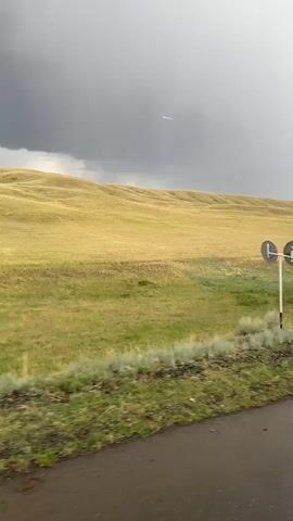 This media file captures a view from a vehicle driving on a paved road through a vast, rolling landscape. The scene is characterized by fields of dry, golden grass that transition to patches of green vegetation closer to the roadside. The ground appears to be sloped, creating undulating hills that stretch towards the horizon. The sky is overcast with heavy, dark clouds, suggesting impending or ongoing rainfall. The road surface is wet, indicating recent precipitation.

The primary subject is the natural scenery, emphasizing the expansive grasslands and the dramatic weather. There are no people or animals visible. A single road sign, a white triangle with black symbols, is present on the right side of the frame, hinting at a rural or undeveloped location. The overall mood is somber and somewhat desolate due to the stormy weather and the barren, wide-open terrain. The time of day is difficult to determine precisely due to the thick cloud cover, but the light appears diffused. The location is identified as Bürkitti, Kazakhstan, which aligns with the description of a vast grassland steppe environment.