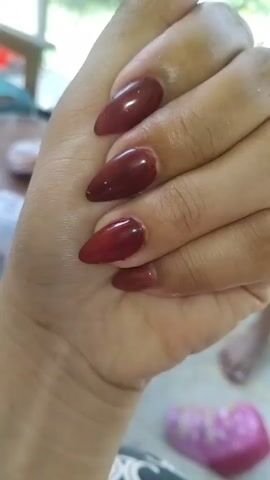 A close-up, high-angle shot shows a left hand with almond-shaped fingernails painted a dark red color. The hand is positioned in the center of the frame, with the fingers curled inward. The background is blurred, but appears to be an outdoor setting with some greenery and furniture visible. There are also hints of other people and objects in the background, including what looks like a pair of feet and a pink object.