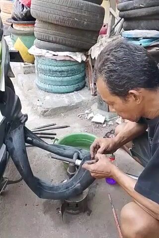 A man with short, dark hair and wearing a black t-shirt is repairing a tire inner tube. He is crouched down on the ground, holding the inner tube in his hands and working on it with tools. The inner tube is black and appears to be for a motorcycle tire. The man is in an outdoor setting, with stacks of tires visible in the background. There is also a green bucket, a metal stand, and various tools on the ground. The man is focused on his work, and his hands are stained with what appears to be grease or dirt. The overall scene suggests a tire repair shop or a roadside mechanic.