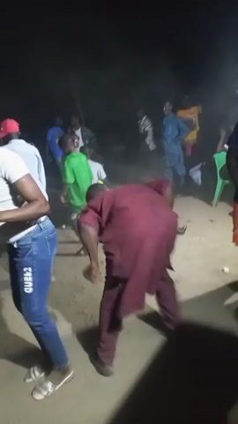 The video shows a group of people dancing at night, apparently at an outdoor party. There are several people visible in the background, some sitting on plastic chairs. One man in a maroon robe is the main focus, dancing energetically. To his left, a person wearing jeans and a white shirt dances along. Another man in a blue shirt and orange shorts joins the main dancer. The ground appears to be sandy. The lighting is dim. The event has a relaxed, casual atmosphere.