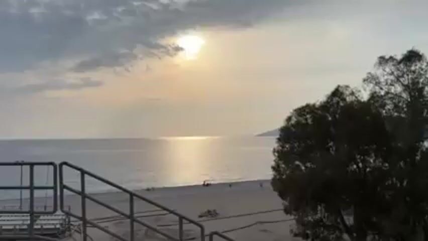 The video presents a view of a beach at what appears to be sunset or sunrise. The sun is partially obscured by clouds, casting a bright glow on the water. The beach is sandy and populated by a few indistinct figures. In the foreground, there is a metal railing suggesting a raised vantage point, possibly stairs or a platform. To the right of the frame, a tree is visible. The overall tone of the scene is serene, with a calm sea and muted colors.