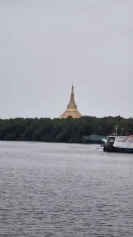 A wide shot shows a golden dome-shaped building behind a line of green trees. In the foreground, there is a body of water with ripples. A boat is partially visible on the right side of the frame. The sky is overcast. The video is looped.