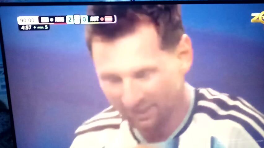 The media file shows a close-up of a soccer game broadcast on a screen. The game is in its final moments, with the score displayed as ARG 2 - 0 AUT, and the time is 90:00 plus 5 minutes of stoppage time. The primary subject is a man, likely a soccer player, with short dark hair and a light complexion, appearing emotional as he is hugged by another player. He is wearing a white and light blue striped jersey with black accents on the sleeves and collar. The background is a blur of green, suggesting a soccer field. The overall mood appears to be one of victory or celebration, given the score and the embrace. The setting is on a screen displaying a sports broadcast, with digital overlays indicating game information. There is no clear indication of weather or time of day within the scene itself, beyond what is displayed on the broadcast graphic. The provided location context of Chirakchi, Uzbekistan, is not visually represented in the media file.
