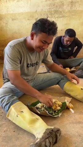 The video shows three men sitting on the floor, eating meals served on banana leaves. The man in the foreground, wearing a grey t-shirt and blue jeans, is smiling and eating with his hands. He is wearing tall yellow rubber boots. To his right, another man in a black long-sleeved shirt and green pants is also eating. A third man, in a brown t-shirt and dark pants, is sitting further away in the background and eating with a utensil. He is wearing flip-flops. The room has a rough, yellow-ochre wall and a concrete floor. A window is visible in the background of the third man's shot. The lighting appears to be natural, coming from the window. The overall impression is of a casual meal break.