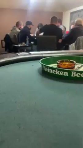 The scene shows a group of approximately eight people sitting around a poker table, engaged in a card game.  The view is from across the table, emphasizing a green Heineken beer tray with a filled ashtray on it in the foreground.  The players are out of focus but appear to be mostly men, with one possibly a woman.  They are dressed in casual clothing. Some are looking at their cards, others are interacting. The room is dimly lit, with a light source overhead.
