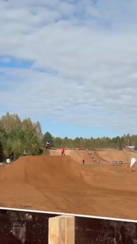 A motocross race is underway on a dirt track under a cloudy sky. Several riders on dirt bikes navigate a series of jumps and turns. The track is made of red dirt and is lined with trees and flags. Some spectators are visible in the distance.