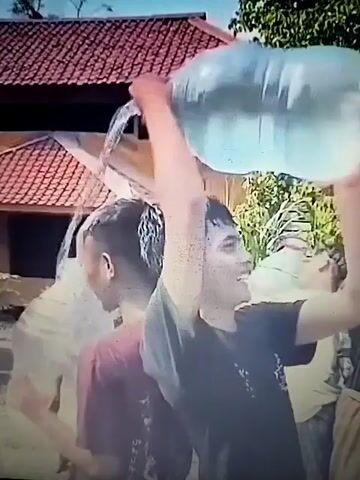 A young man pours water from a large plastic container onto the head of another young man. The recipient is wearing a maroon shirt and has short, dark hair that is slicked back and wet from the water. The person pouring the water is wearing a dark t-shirt and is smiling. They are outdoors, with lush green foliage in the background. In the foreground, another person wearing a white shirt and dark pants is visible, bent over. It appears to be a hot day, possibly a celebration or a playful activity.