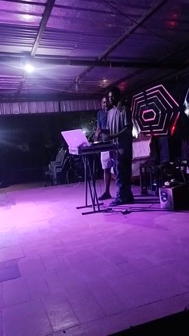 The video captures a scene inside what appears to be a makeshift or temporary structure, possibly for an event or performance. Two individuals are standing near a table laden with equipment, including a laptop, which suggests they might be involved in sound mixing or DJing. Above them is a hexagon-shaped light fixture emitting a reddish-pink glow. The background is a mix of architectural elements, with metallic frames and translucent panels. The lighting is predominantly blue and purple, adding to the ambient mood.