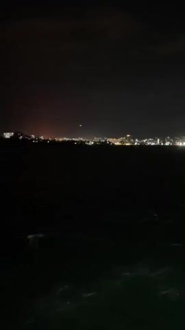 The video shows a nighttime cityscape across a dark body of water. The city lights are white and blurry, forming a line along the horizon. There are occasional orange lights as well. The water in the foreground is black, with small white waves visible. The sky is dark gray. There are muffled voices in the background.