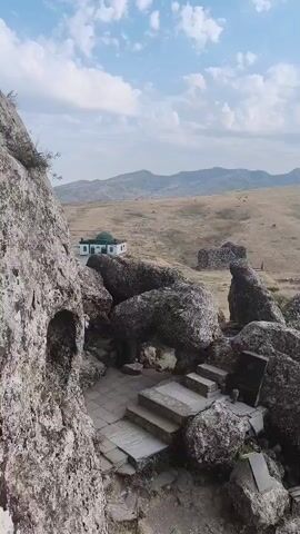 A drone flies over a rocky, barren landscape, showing a small white building with a green dome in the distance. In the foreground, large boulders are scattered around a set of stone steps leading up to an opening in a rock face. The sky is blue with scattered clouds.