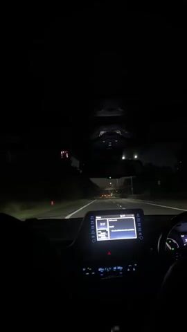 The video shows the interior of a car at night, looking forward through the windshield. The dashboard is visible with a touchscreen display showing a song title, and buttons. The car is on a highway, with other cars visible in the distance. Streetlights and trees line the road. The view from inside the car suggests the person is a passenger or the driver is recording.