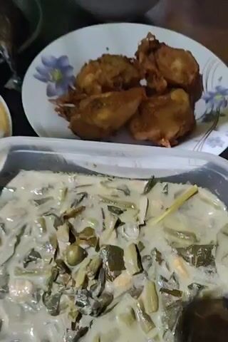 The video shows a table set with a variety of Filipino dishes. In the foreground is a clear rectangular container filled with vegetables cooked in coconut milk, likely a dish called "ginataang gulay". Above this, there is a white plate with blue floral patterns, holding several pieces of what appears to be fried banana or a similar sweet treat. Moving to the right, there is a plate containing fried fish, specifically "bangus" or milkfish. Below the fish is a plate of chopped tomatoes and onions, a common condiment known as "kinilaw" or "ensalada". The table itself has a simple, neutral-colored surface, and the environment seems to be indoors.