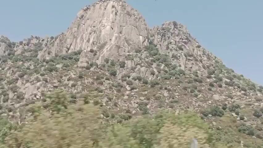 A video shows a mountain with a clear sky. The mountain is rocky with patches of green vegetation. Some birds can be seen flying in the sky above the mountain. The camera appears to be moving, as evidenced by slight shifts in perspective and the appearance of a partial, blurry object in the foreground at the beginning of the video. The overall impression is of a scenic outdoor view.