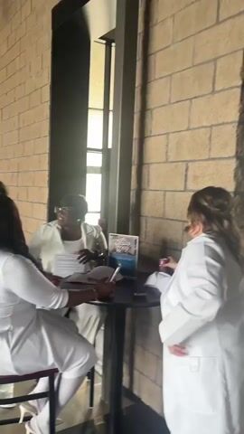 The video captures a group of six women gathered in what appears to be an indoor event space or lobby, characterized by a light brown brick-patterned wall and modern architecture. All the women are dressed in white or off-white attire, suggesting a themed event or uniform.

In the foreground, a woman with long dark hair is seated at a high-top table, seemingly engaged with papers or a device placed on it. To her left, another woman in a white dress, white tights, and white shoes holds a phone, possibly capturing the moment. Behind the seated woman, a third woman, also in white pants and a jacket, is smiling and interacting. Two additional women in white are visible in the background, with another partially visible on the far right.

The setting includes a tall, dark-framed structure that resembles a window or mirror, reflecting parts of the room and bright light from outside. A plant is visible in the background to the left. On the far left wall, a sign with the text "Preschool" is partially visible. Further along the brick wall, a white poster features the stylized text "Love People." On the table, various items are present, including papers, possibly a laptop or tablet, and a brochure or book with the visible text "The City..." and potentially "Connection" underneath a cityscape image. The women appear engaged and happy, suggesting a social or celebratory atmosphere.