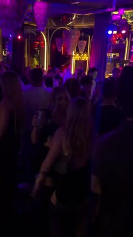 A crowded nightclub is illuminated by purple and pink lights. People are dancing and socializing. In the foreground, two women are visible, one with blonde hair and the other with brown hair, both wearing black outfits. The woman with blonde hair is holding a drink. The background shows a DJ booth and other club patrons.