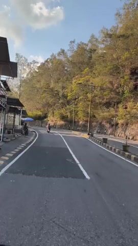 A motorcycle is riding down a paved road that curves to the left. The road is bordered on the left by a sidewalk with a small food stall and a person sitting under a blue umbrella. On the right, the road is bordered by a rocky slope covered in trees. Streetlights are visible along the right side of the road. The sky is blue with some clouds. The sun is shining, casting shadows. The overall scene depicts a scenic road with a lone rider and a small roadside vendor.