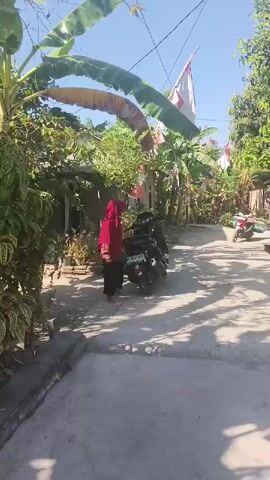 A person wearing a red hijab and dark pants is walking and gesturing in a street. There are motorcycles parked along the side of the street and trees and buildings in the background. There are also Indonesian flags hanging in the background.