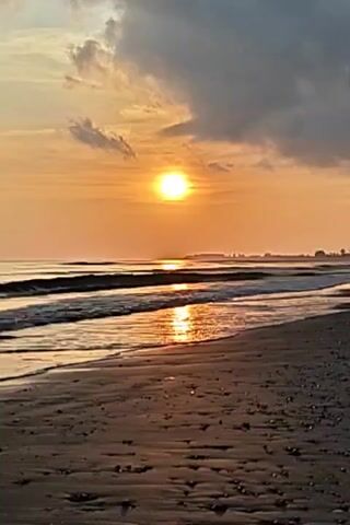The scene depicts a beach at sunset. The sun, appearing as a bright white orb against an orange sky, is partially obscured by clouds. The waves gently roll onto the shore, reflecting the sunlight on the water's surface. The beach itself is sandy and appears slightly wet from the receding tide. In the distance, a stretch of land with some trees or structures can be seen on the horizon.