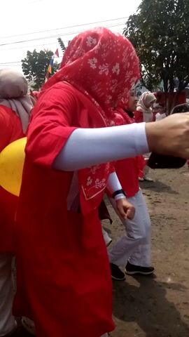 This video shows a group of people, mostly women, dressed in matching red outfits with white headscarves and white pants. They appear to be participating in a lively outdoor activity, possibly a dance or a game, given their synchronized movements and joyful expressions.

The scene is set in Sukaraja, Indonesia, with glimpses of trees, buildings, and what looks like a street or paved area in the background. The time of day appears to be daytime, with bright, natural light suggesting fair weather. The overall atmosphere is energetic and festive, with participants smiling and engaged in the activity.

The subjects are a group of individuals, likely performing in some sort of cultural or community event. The primary objects visible are the matching red clothing and white headscarves. The setting is outdoors, with some indication of a public space or venue.

Notable details include the coordinated red attire, suggesting a team or group identity. The people are in various dynamic poses, indicating movement and activity. The background offers subtle cues about the location, such as architectural styles and vegetation typical of Indonesia. The emotions conveyed are predominantly positive, with smiles and animated body language.

There is no discernible text visible in the video.