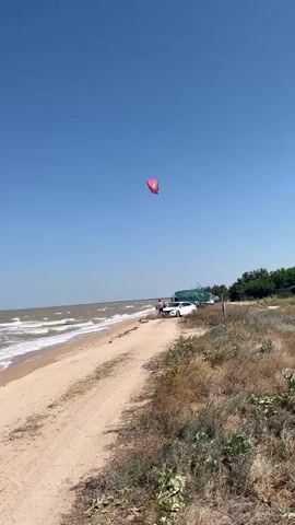 The video shows a beach on a clear day. The sea has moderate waves. There is a sandy path along the beach, with dry grass and shrubs on one side. A red kite is visible in the sky. A white car and two people are standing near the car on the path.