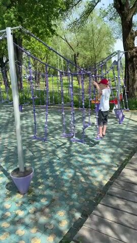 The video shows a young boy navigating a purple rope climbing structure in a park. The boy, wearing a red baseball cap, a light-colored t-shirt, and dark shorts, carefully steps across the rope ladders while holding onto the ropes above. The structure is made of purple ropes and wooden steps. The ground underneath is covered with blue and green patterned tiles. Trees surround the play area, and sunlight casts shadows on the ground.