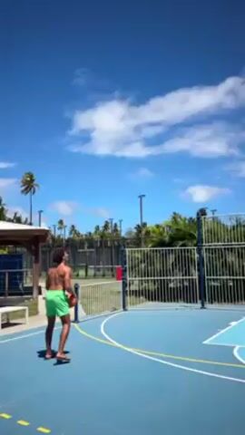 A short video shot outdoors on a sunny day shows a person playing basketball. A person wearing green shorts is standing on a blue basketball court, facing away from the camera. They are in a shooting stance with a basketball held to the side. They shoot the basketball towards the basketball hoop. The basketball goes into the basket. The person then walks away. Tall palm trees and lush greenery are visible in the background, along with a white fence around the basketball court. There are also a few small buildings and light posts in the background. The sky is blue with fluffy white clouds.