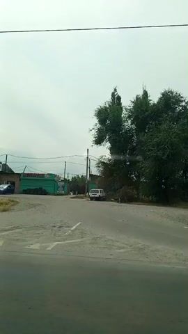 The video shows a street scene with several cars and buildings. The sky is overcast. On the left side of the road, there is a building with the text "АВТОМОЙКА" which translates to "car wash". There are several cars parked in front of the building, including a blue sedan and a white SUV. A tall, slender tree stands in front of the building. Further down the road, there is another building with signs that include "ГАЗ" which indicates a gas station. There are more cars parked in front of this building, including a white station wagon and a silver sedan. On the right side of the road, there are more trees and a white car is visible. The road appears to be asphalt. There is a traffic sign visible in the foreground which is a triangle pointing downwards with a red border and a white background.