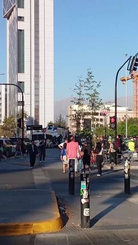 The video shows a city street intersection during the day. A tall, modern skyscraper is visible in the background. There are several people crossing the street, and a few bicyclists are also present. There is traffic on the street, including cars and taxis. A construction crane is visible in the distance. The sky is clear and blue.