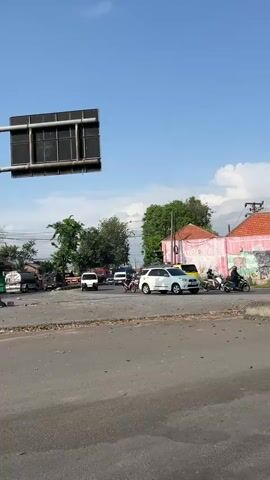 The video shows a sunny outdoor scene with a clear blue sky. The foreground is a paved area, possibly a road or parking lot, with some debris scattered on the ground and concrete barriers. In the mid-ground, there are several vehicles, including a white SUV, a yellow taxi, a motorcycle with a rider, and a few other cars. Some buildings with pink roofs and white walls are visible in the background, along with trees and power lines. A large signpost is partially visible at the top left of the frame. The overall impression is of a busy street intersection or a marketplace.