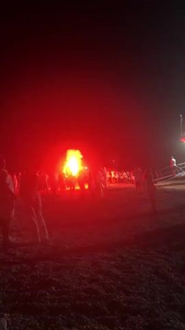 A group of people are gathered on a beach at night around a bonfire. The flames are visible, as is a large cloud of red smoke from a flare. The people are mostly silhouetted against the light of the fire and flare. Some individuals are wearing white clothing. To the right of the frame, a portion of a boat or structure with lights is visible. The ground appears to be sandy or grassy.