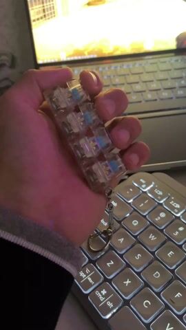 A person's left hand, clad in a grey sleeve, is holding a clear, rectangular, multi-switch device that functions as a keychain. The device contains several individual mechanical switches, each encased in clear plastic. As the person squeezes or presses the switches, they emit clicking sounds and light up in various colors. Initially, some switches are blue, others green. Throughout the video, the lights predominantly glow green, with a brief flash of red from one of the switches. A small metallic chain with a keyring and clasp hangs from one end of the device.

In the background, a silver laptop with a dark keyboard is open, its screen displaying what appears to be a brightly lit game or graphic, predominantly in yellow and orange hues. To the right and foreground of the laptop is a separate, external keyboard, likely a wireless one, with square, backlit keys. The keycaps are illuminated, showing letters such as 'Q', 'W', 'E', 'R', 'A', 'S', 'D', 'F', 'Z', 'X', 'C', 'V', and function keys like 'esc', 'tab', 'caps lock', 'opt', 'start', 'cmd', and 'alt'. The overall setting suggests an indoor environment, possibly a desk, with ambient lighting allowing the illuminated devices to stand out prominently. The focus is on the interactive, tactile, and visual elements of the switch-tester keychain.