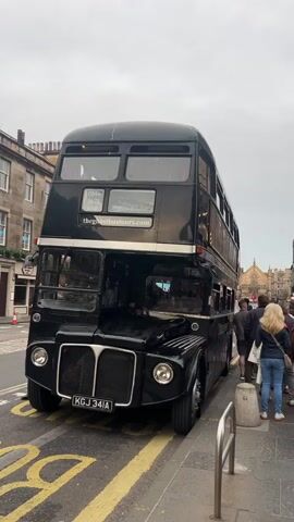 A vintage black double-decker bus is parked on a street in front of a building. The bus has two floors and the license plate reads "KGJ 341A". A group of people are standing on the sidewalk to the right of the bus, some of them are looking at the bus. The street in front of the bus has yellow lines painted on it. The sky is overcast.