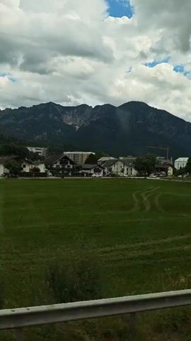 The video shows a view from a vehicle moving alongside a grassy field. In the distance, there are houses and buildings, and behind them, a large mountain range under a cloudy sky. Trees line the side of the road, with some foliage visible in the foreground. There are no people or animals clearly visible. The lighting suggests it is daytime.