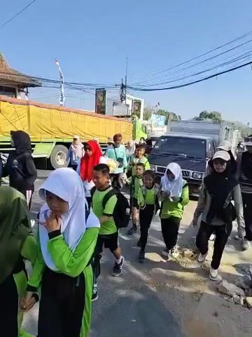 A group of people, many of whom appear to be children wearing green shirts and black pants, are walking along a road. Some of the children are wearing white hijabs. There are adults accompanying them, also wearing hijabs and modest clothing. In the background, there is a large yellow truck, a smaller black truck, and a few other vehicles and people. The sky is blue and clear. There are power lines visible overhead. The ground appears to be a paved road with some debris on the side. Some individuals are holding water bottles. The group seems to be moving in the same direction.