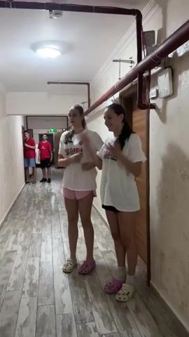 This video captures two teenage girls standing in a hallway, dressed in white t-shirts with "BULGARIA" printed on them in red and green, and pink shorts. They are wearing Crocs and appear to be engaged in a synchronized dance or cheer. Their movements are energetic and playful.

In the background, further down the hallway, three teenage boys are visible. They are wearing red t-shirts and shorts, and also seem to be part of a group activity, possibly cheering or participating in a competition. One boy is looking directly at the camera.

The setting appears to be a dormitory or a similar residential building, indicated by the long hallway, the textured walls, the overhead lighting, and the visible pipes and electrical conduits running along the ceiling. There are doors visible on the right side of the hallway, one of which has a number "112" on it. The floor is tiled with a distressed wood-look pattern.

The lighting suggests it is daytime, though there are no windows visible to confirm the exact time of day or weather conditions. The overall mood seems positive and lively, with the subjects appearing enthusiastic and engaged in their activity. The presence of the Bulgarian national colors and text on the t-shirts suggests they might be participating in a sporting event or cultural exchange related to Bulgaria, possibly in Çanakkale, Turkey, given the location context.