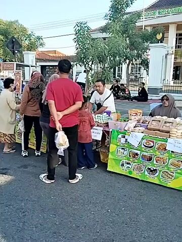 The video captures an outdoor market scene from a fixed perspective, panning right to left. People are gathered around various stalls, some sitting on the ground, others standing. Several food stalls are visible, offering items like "JASUKE JAGUNG MOZZA MOLORR" and "PECEL LELE." Vendors, predominantly women, are attending to customers. The environment is bustling with activity, suggesting a lively market atmosphere. In the background, a large building with a sign that reads "IKATAN HAKIM INDONESIA (IKAHI)" is visible behind a fence. The lighting indicates it's daytime, possibly morning or late afternoon.