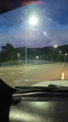 The video appears to be captured from inside a vehicle moving forward on a road. The time is likely dusk or early evening, as the sky is dark blue with some clouds, and streetlights are illuminated. On the right side of the road, there are several streetlights lining the pavement. On the left side, there is a bridge with a metal railing. In the distance, there are hills or mountains, and a few buildings are visible on the slopes. The interior of the car is partially visible in the foreground, showing part of the dashboard and windshield wipers. The video is 5 seconds long.